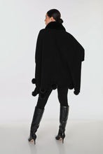 Load image into Gallery viewer, A woman shown from the back wearing a black poncho with a faux fur collar and pom-pom trim, paired with black leggings and black knee-high boots, against a light gray studio background.
