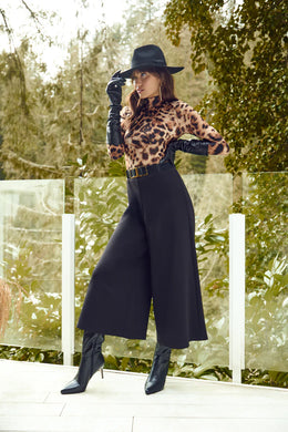 Woman in leopard print top, black wide-leg pants, black hat, gloves and boots posing outdoors.