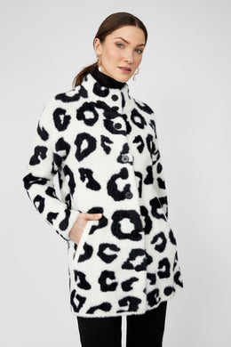 Woman wearing a white coat with black leopard print pattern, black pants and gold earrings against a white background.