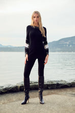 Load image into Gallery viewer, Woman wearing a long sweater with white and black patterned sleeves with crystal embellishments, black pants and tall black boots standing in front of a body of water with hills in background.
