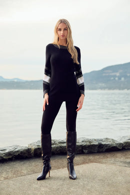 Woman wearing a long sweater with white and black patterned sleeves with crystal embellishments, black pants and tall black boots standing in front of a body of water with hills in background.
