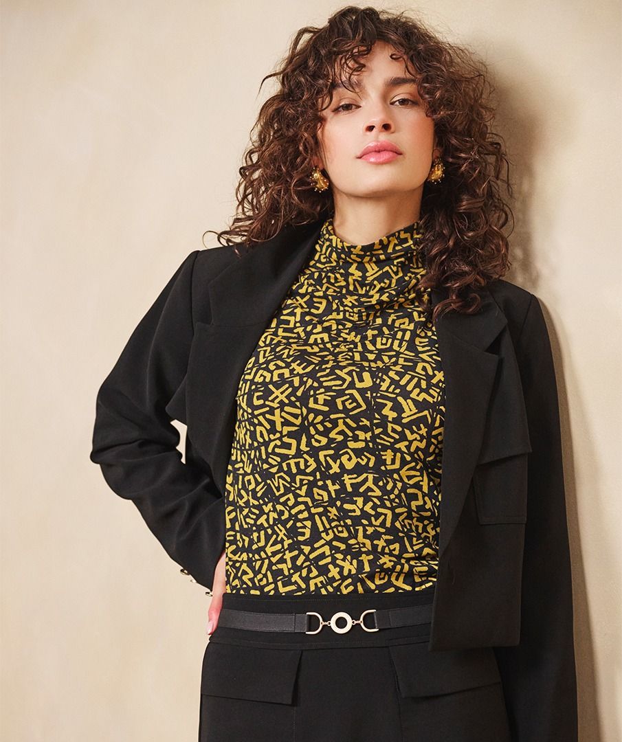 Woman wearing a black blazer and yellow patterned shirt against a beige background.