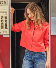 Load image into Gallery viewer, Woman in a red shirt and blue jeans standing inside a door with a poster on the wall.
