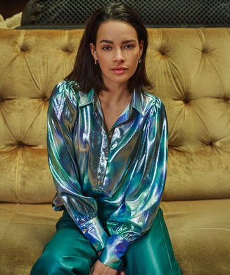Woman wearing a shiny teal, blue and silver blouse and teal faux leather pants sitting on a beige couch.
