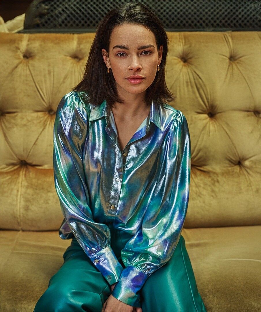 Woman wearing a shiny teal, blue and silver blouse and teal faux leather pants sitting on a beige couch.
