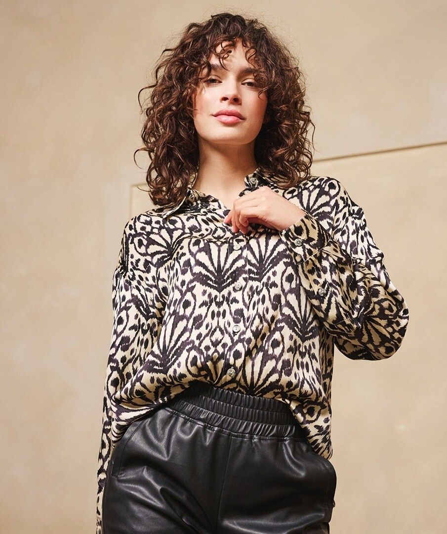 Woman wearing an abstract print top in black and beige and black pants against a beige background.