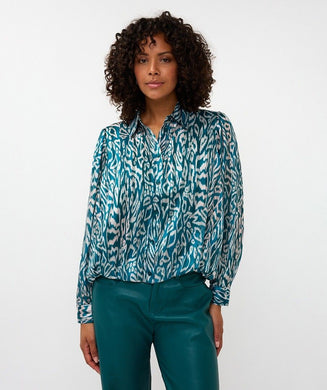 Woman wearing a teal and white with shimmer patterned blouse and teal pants against a white background.