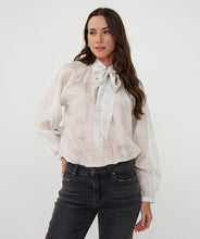 Load image into Gallery viewer, Woman wearing an off-white blouse with a tie detail and dark jeans against a white background.
