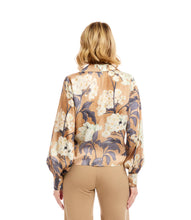 Load image into Gallery viewer, Woman wearing a tie waist patterned long sleeve blouse in floral grey, cream and tan and tan pants against a neutral background.
