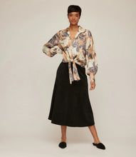 Load image into Gallery viewer, Woman wearing a tie waist patterned long sleeve blouse in floral grey, cream and tan, long black skirt and black shoes against a neutral background.
