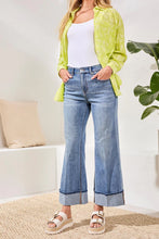 Load image into Gallery viewer, Woman wearing a bright green top over a white top, blue jeans and white sandals standing in a room with a plant and pillow in background.
