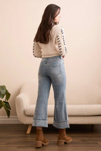Load image into Gallery viewer, Woman wearing a patterned sweater, palazzo jeans, and brown boots standing in a room with a beige sofa and plant.
