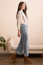 Load image into Gallery viewer, Woman wearing a patterned sweater, palazzo jeans, and brown boots standing in a room with a beige sofa and plant.
