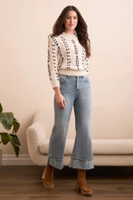 Load image into Gallery viewer, Woman wearing a patterned sweater, palazzo jeans, and brown boots standing in a room with a beige sofa and plant.
