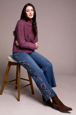 Woman sitting on a stool wearing a purple sweater, blue jeans with floral embroidery and black boots against a neutral background.