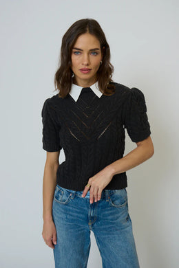 Woman wearing a black knit top with a white collar and blue jeans against a plain background.