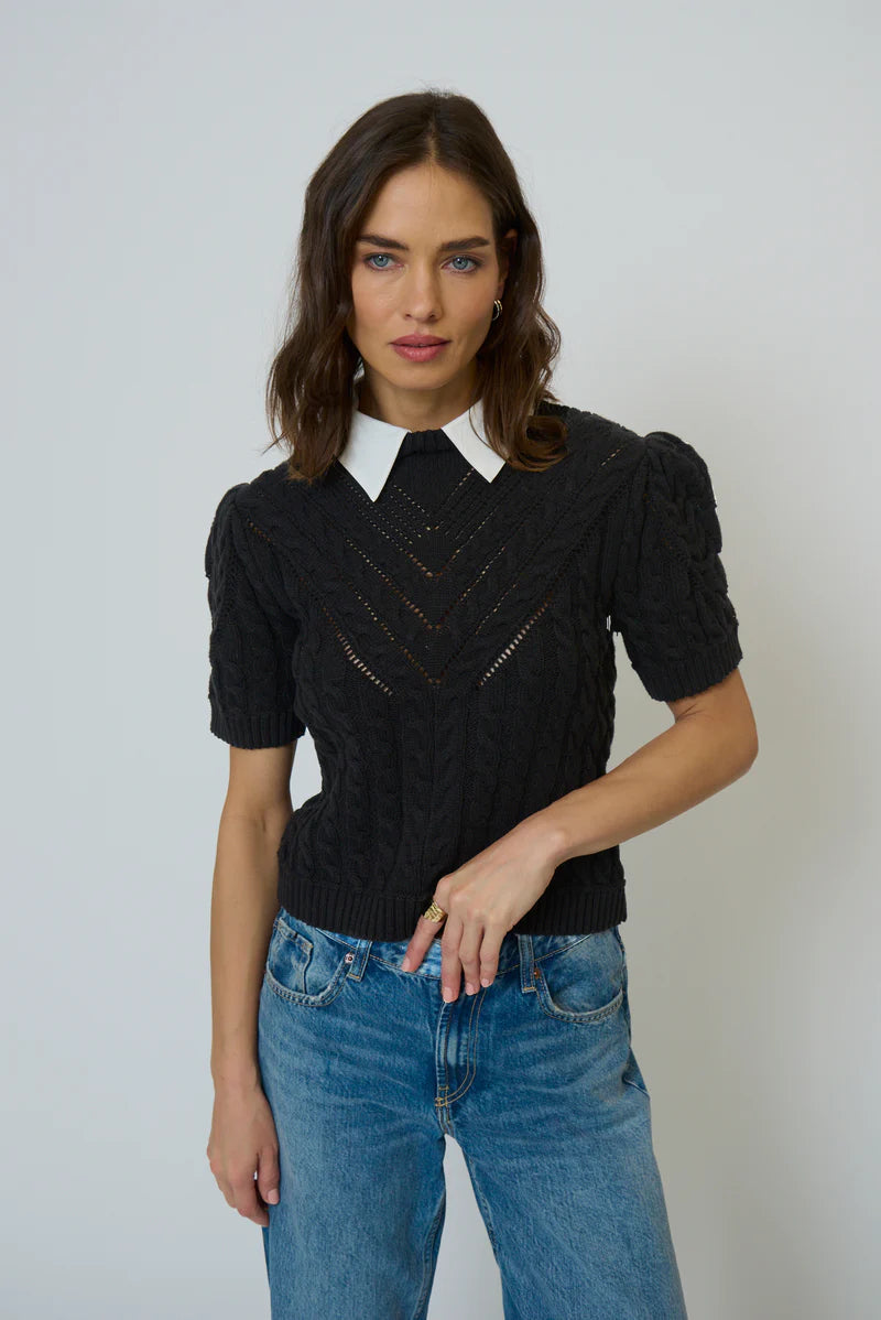 Woman wearing a black knit top with a white collar and blue jeans against a plain background.