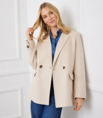 Woman wearing an oatmeal overcoat over a blue shirt and blue jeans against a white wall.