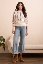 Load image into Gallery viewer, Woman wearing a cream sweater with black crochet stitching throughout, light blue jeans, and brown boots standing in a room with a beige sofa and plant.
