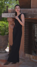 Load image into Gallery viewer, Woman in a black sleeveless jumpsuit standing in front of a stone wall with greenery in the background.
