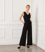 Load image into Gallery viewer, Woman wearing a black sleeveless jumpsuit against a white paneled wall.
