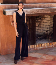 Load image into Gallery viewer, Woman in a black sleeveless jumpsuit standing in front of a brick fireplace.
