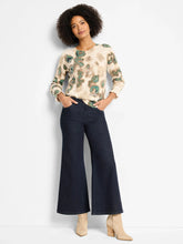 Load image into Gallery viewer, Woman wearing a beige and metallic sweater with green camo pattern, wide leg blue jeans and tan suede boots against a plain background.
