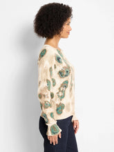 Load image into Gallery viewer, Woman wearing a beige and metallic sweater with green camo pattern and blue jeans against a plain background.
