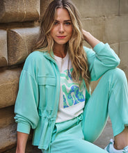 Load image into Gallery viewer, Woman in mint green tracksuit sitting against a stone wall.

