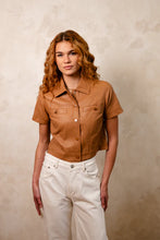 Load image into Gallery viewer, Woman wearing a short sleeve brown leather shirt and white pants against a beige background.
