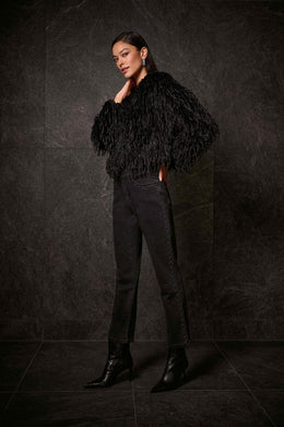 Woman wearing a black faux fur jacket, dark grey studded jeans and black boots against a dark wall.