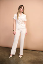 Load image into Gallery viewer, Woman wearing an off-white short sleeve draped blouse and off-white pants with off-white heels against a dark beige wall.
