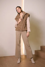 Load image into Gallery viewer, Woman wearing a beige puffer vest over a beige top, warm taupe wide-leg jeans and beige suede boots standing next to stairs in a minimal indoor setting.
