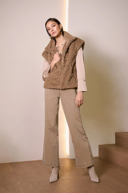 Woman wearing a beige puffer vest over a beige top, warm taupe wide-leg jeans and beige suede boots standing next to stairs in a minimal indoor setting.