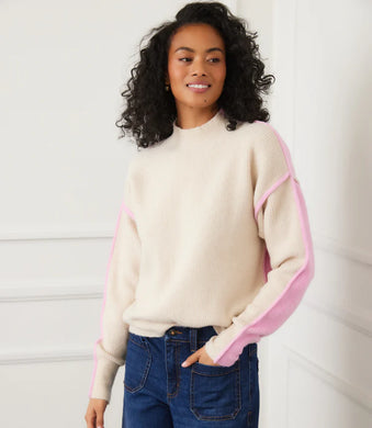 Woman wearing a sand and pink colorblock sweater and jeans against a white wall.