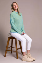 Load image into Gallery viewer, Woman sitting on a wooden stool wearing a teal blue sweater, white leggings against a pale pink background.
