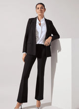 Load image into Gallery viewer, A woman models a black pantsuit consisting of a tailored blazer and matching trousers, styled with a white button-down shirt and clear high-heeled sandals, set against a minimalist white studio background.
