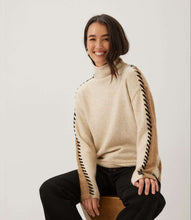 Load image into Gallery viewer, Woman wearing a color block turtleneck sweater with black contrast stitching down the arms and black pants sitting on a stool against a white background.

