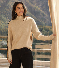 Load image into Gallery viewer, A woman wearing a color block, turtleneck sweater with black stitching down the arms, paired with black pants, standing by a window with a mountain landscape in the background.

