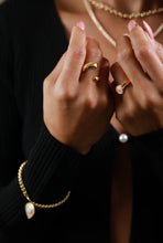 Load image into Gallery viewer, Close-up of a person wearing multiple gold rings and a gold chain bracelet with a teardrop moonstone pendant, paired with a black ribbed sweater and layered gold necklaces.
