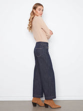 Load image into Gallery viewer, A woman stands in profile wearing a beige ribbed knit mock-neck top, dark blue wide-leg cropped jeans, and tan heeled ankle boots against a plain white wall.
