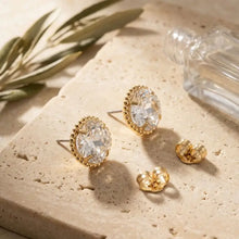Load image into Gallery viewer, Gold earrings with clear gemstones on a textured surface with a plant and glass container in the background.
