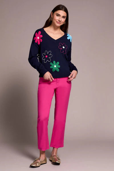 Woman wearing a navy sweater with colorful flower patterns, bright pink pants and metallic flats against a neutral background.