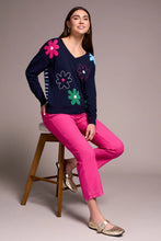 Load image into Gallery viewer, Woman wearing a navy sweater with colorful flower patterns, bright pink pants and metallic flats sitting on a bench against a neutral background.
