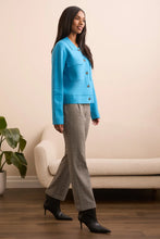 Load image into Gallery viewer, Woman wearing a blue jacket, gray pants and black boots standing in a room with a beige sofa and plant.
