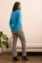 Load image into Gallery viewer, Woman wearing a blue jacket, gray pants and black boots standing in a room with a beige sofa and plant.
