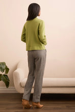 Load image into Gallery viewer, Woman wearing a green jacket, gray pants and brown boots standing in a room with a beige sofa and plant.
