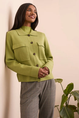 Woman wearing a green jacket and gray pants standing against a light pink wall with a plant to the right.