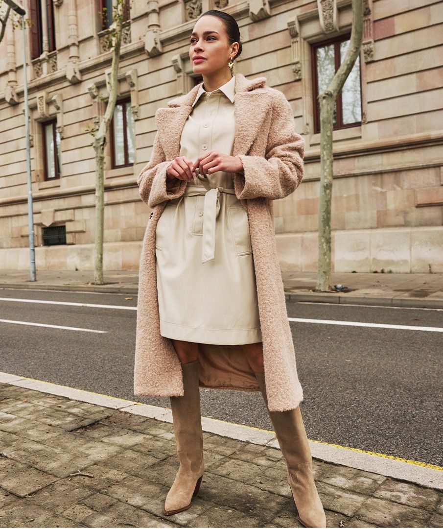 Woman wearing a long beige coat over a light sand faux leather dress and beige suede boots standing on a street with a building in the background.