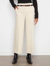 Load image into Gallery viewer, A person wearing high-waisted, wide-leg cropped trousers in a beige color, styled with a dark brown belt, a black ribbed long-sleeve top, and black ankle boots against a white wall.
